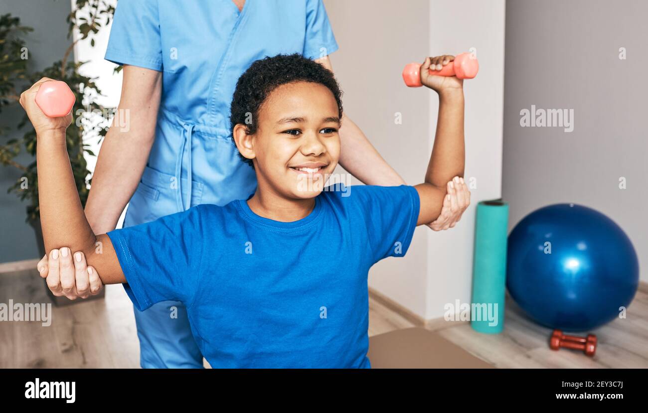Afroamerikanischer Junge trainiert mit Physiotherapeuten mit Kurzhanteln im Reha-Zentrum. Lächelndes Kind mit den Händen nach oben, mit Hanteln Stockfoto