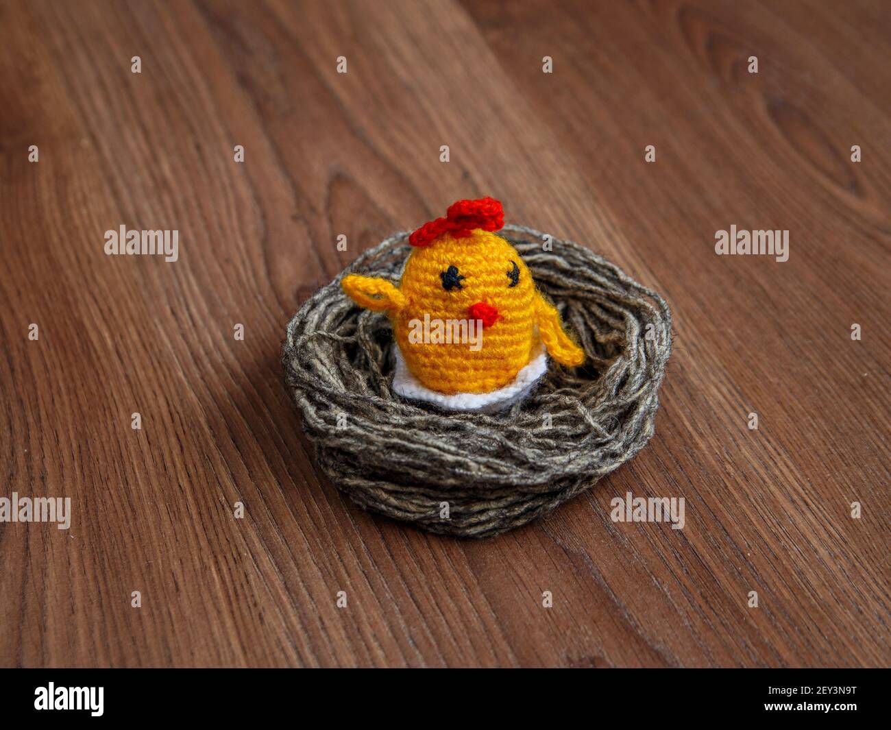 Gelbes Spielzeug-Huhn im Nest aus grauen Wollfäden auf brauner Oberfläche. Stockfoto