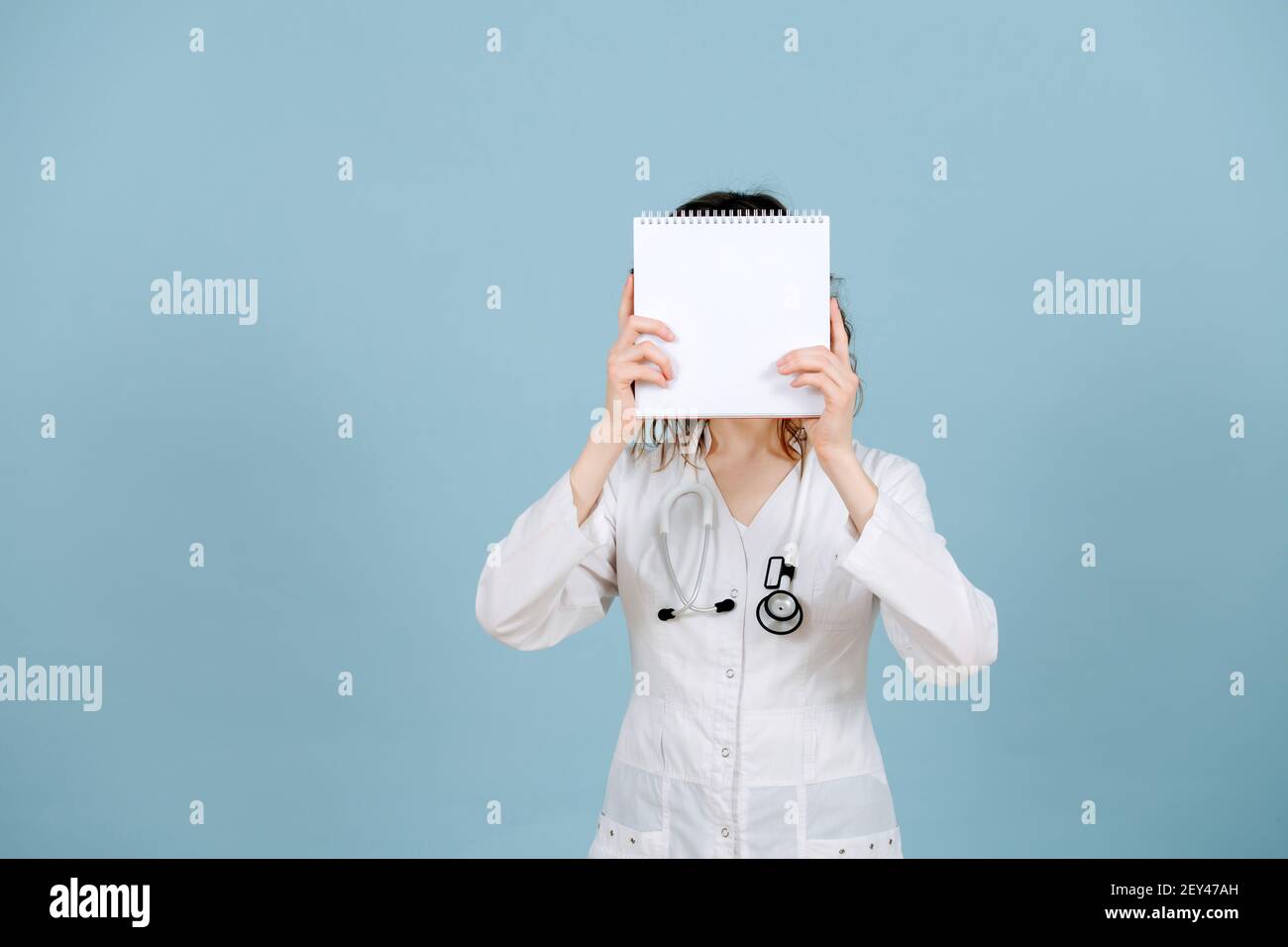 Krankenschwester Frau stehend mit leerem Notizbuch in den Händen, zeigt es. Auf blauem Hintergrund. Sie trägt weiße Robe und Stethoskop. Stockfoto