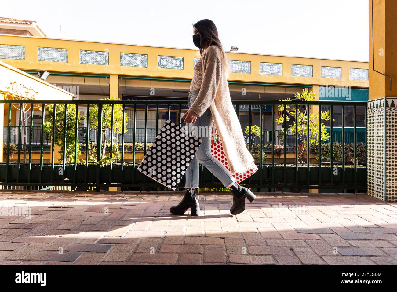 Spanische Brünette Mädchen mit Gesichtsmaske zu Fuß mit Einkaufstaschen in einem Einkaufszentrum. Stockfoto