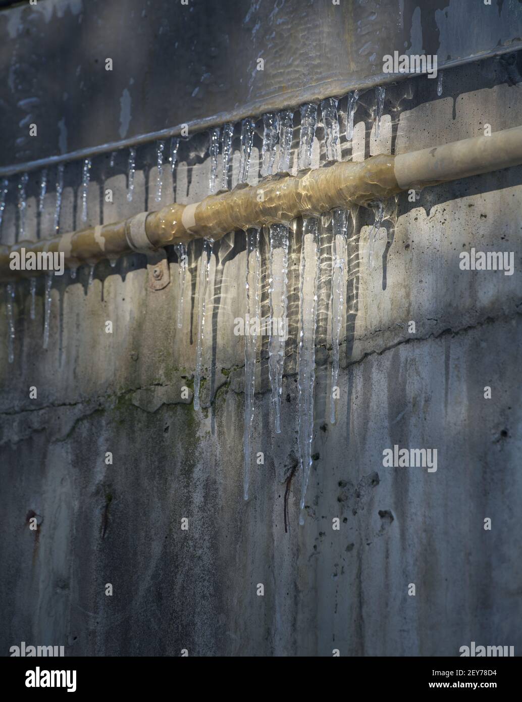 Gefrorene spitzen Eiszapfen, die an einem industriellen Metallrohr auf grunbendem Wasser hängen, beschädigten im Hintergrund Eisschatten auf der Betonaußenwand Stockfoto