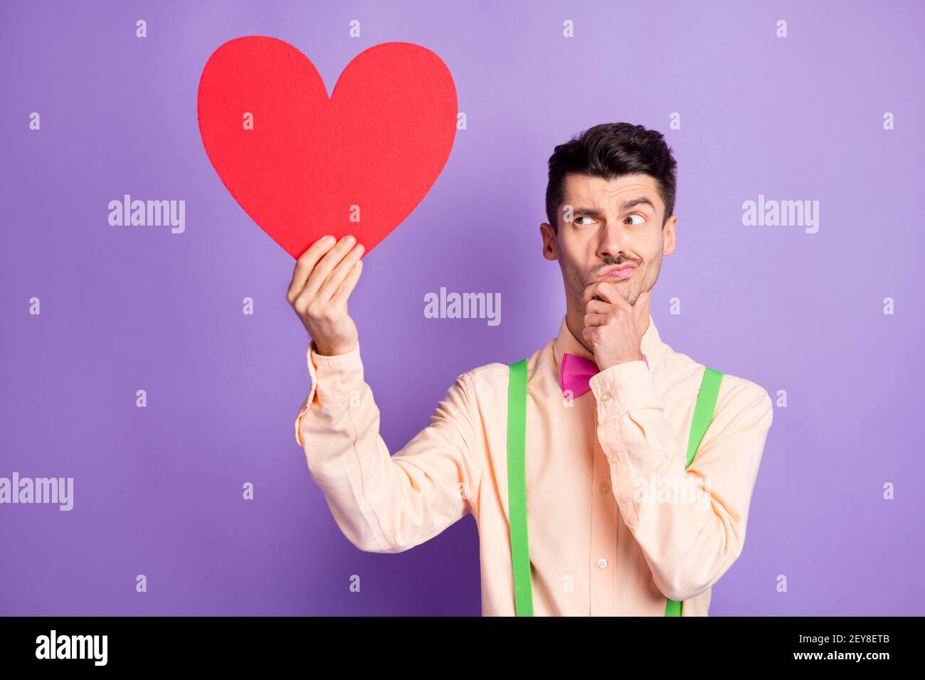 Foto von nachdenklichen unsicher jungen Mann gekleidet gelben Hemd halten Große rote Papier Herz Arm Kinn isoliert lila Farbe Hintergrund Stockfoto
