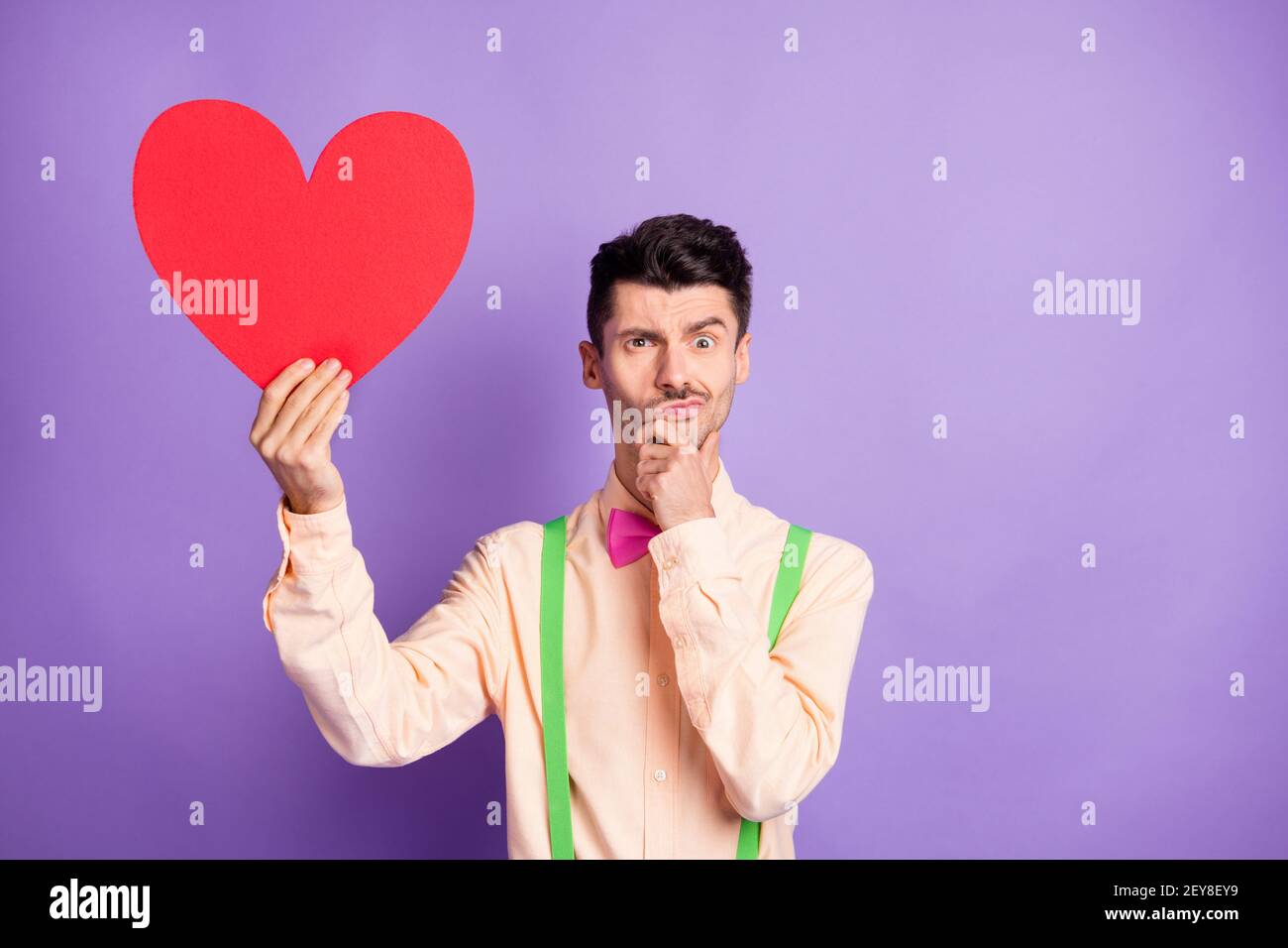 Foto von zweifelhaft ernst jungen Kerl tragen gelbe Hemd Arm Kinn hält große Papier rot Herz isoliert violette Farbe Hintergrund Stockfoto