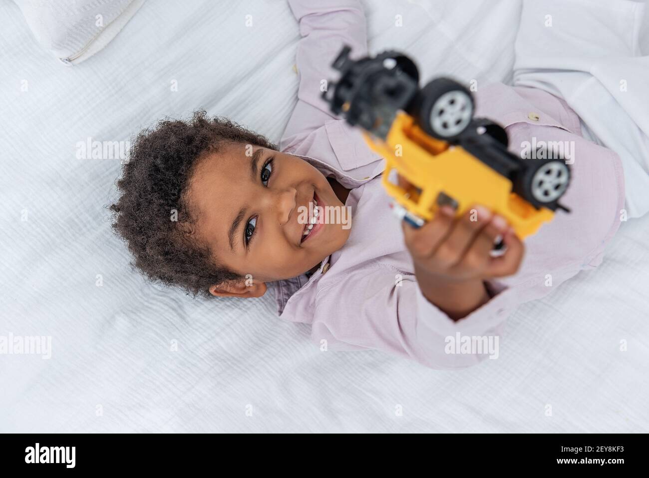 Kleiner afroamerikanischer Junge, der mit Spielzeugautos auf einem weißen Bett auf einem Holzboden spielt. Minimalistisches Interieur, Pampagras, Treibholz an der Wand, luftiger Doo Stockfoto