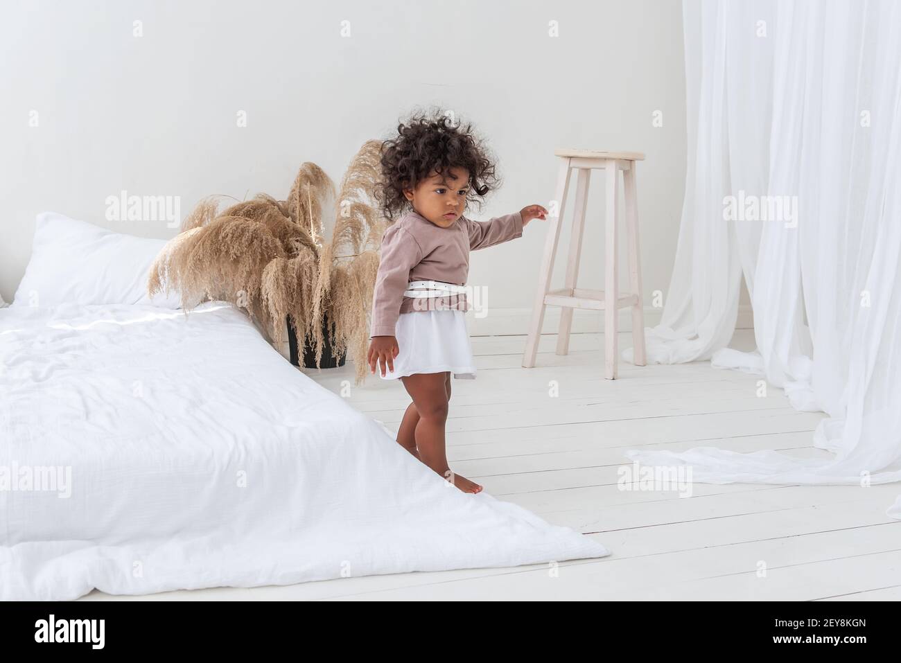 Kleine rothaarige afroamerikanische Mädchen spielt, läuft am weißen Bett, hat Spaß, lacht, lächelt. Das Konzept der Annahme multi-ethnischen Familie. Scandina Stockfoto