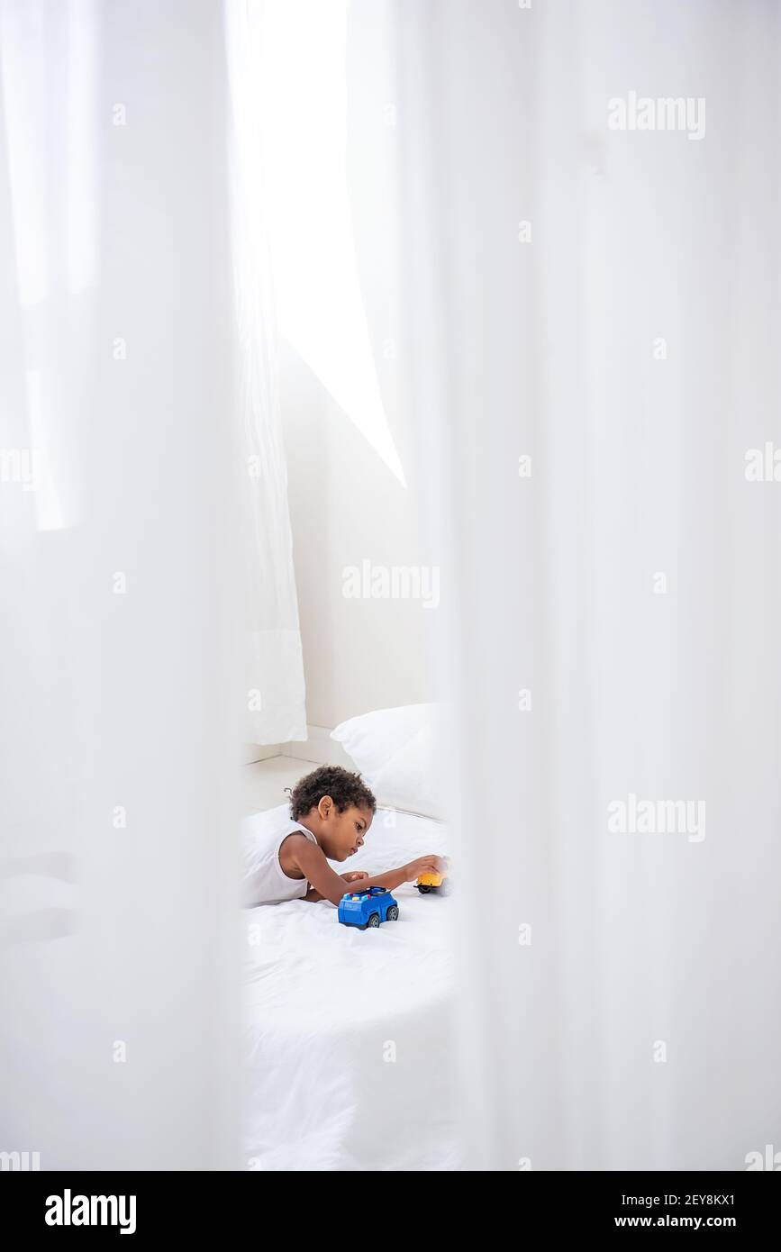 Kleiner afroamerikanischer Junge, der mit Spielzeugautos auf einem weißen Bett auf einem Holzboden spielt. Minimalistisches Interieur, Pampagras, Treibholz an der Wand, luftiger Doo Stockfoto