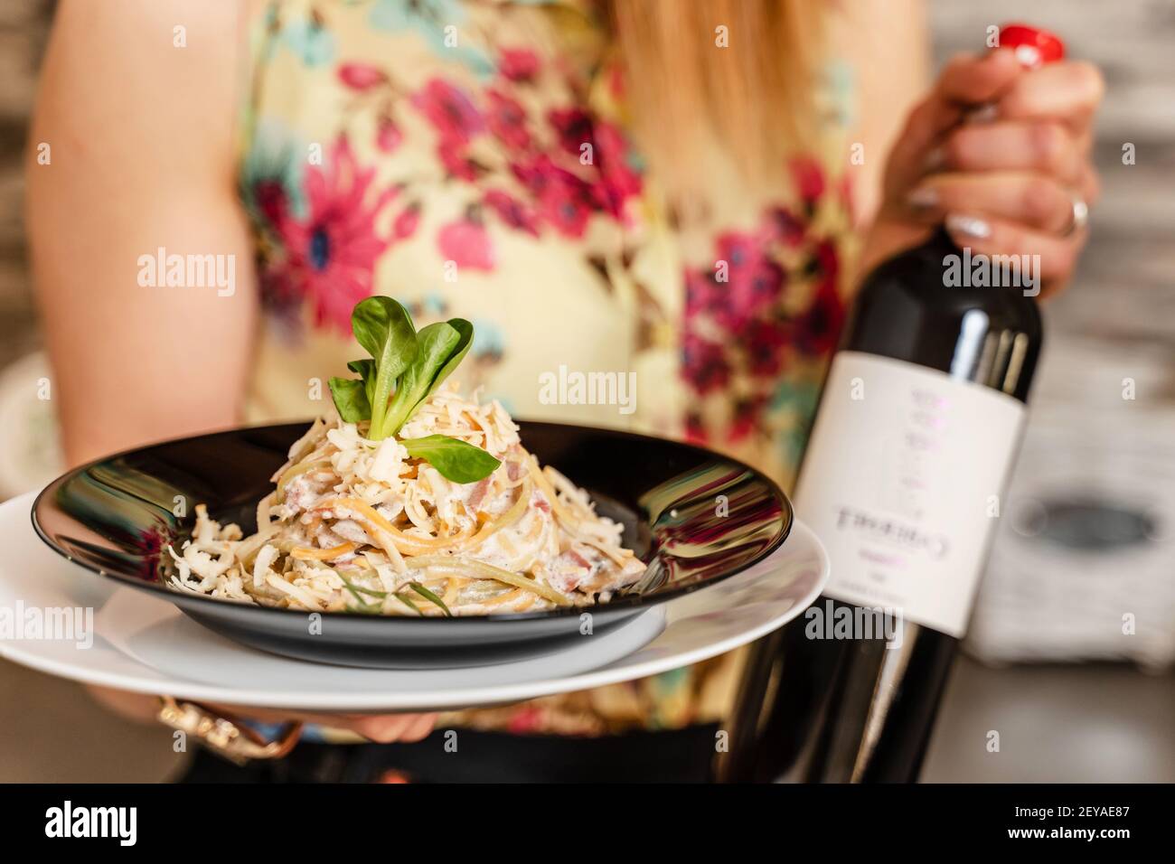 Nahaufnahme des mittleren Abschnitts einer unbekannten kaukasischen Frau mit einem Teller gekochte Carbonara Pasta Spaghetti in einem Teller mit Flasche Wein neben zu Hause - Stockfoto