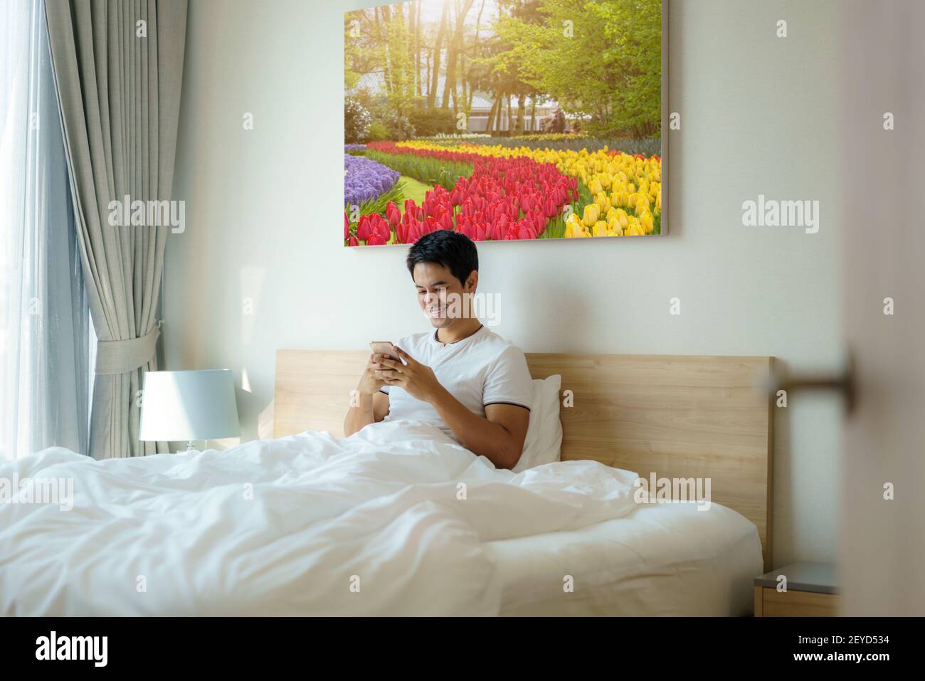 Ein asiatischer Mann sitzt mit einer weißen Decke im Bett und checkt seine E-Mails am Morgen zu Hause auf seinem Handy, bevor er duscht. Stockfoto