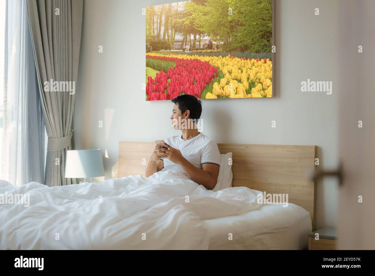 Ein asiatischer Mann sitzt mit einer weißen Decke im Bett und checkt seine E-Mails am Morgen zu Hause auf seinem Handy, bevor er duscht. Stockfoto