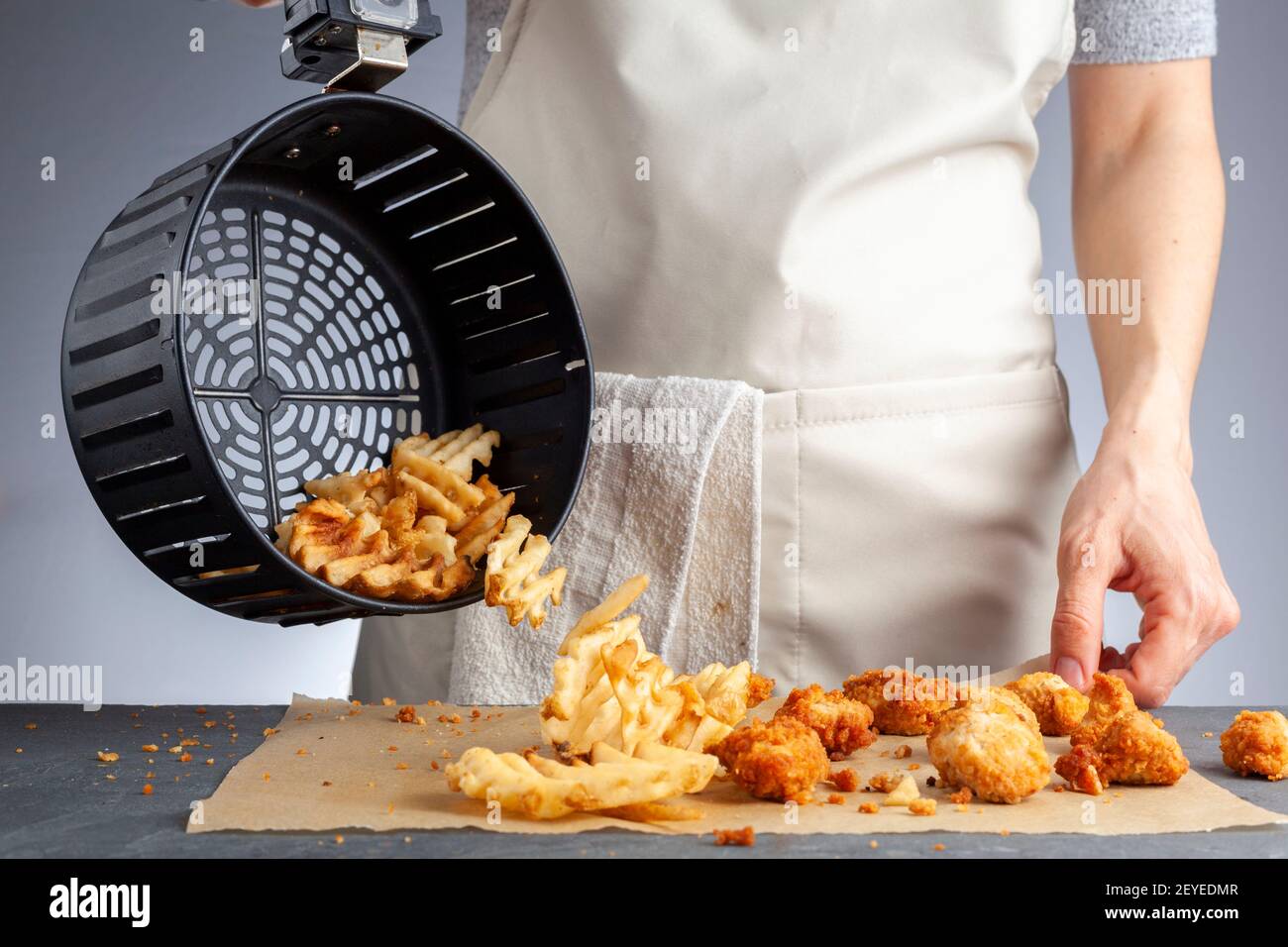 Eine Frau dumpt frisch zubereitete Kartoffelwaffelfritten aus dem Korb zusammen mit Hühnchen-Nuggets auf eine Arbeitsplatte. Sie frittierte sie in der Airfryer mit sehr Stockfoto