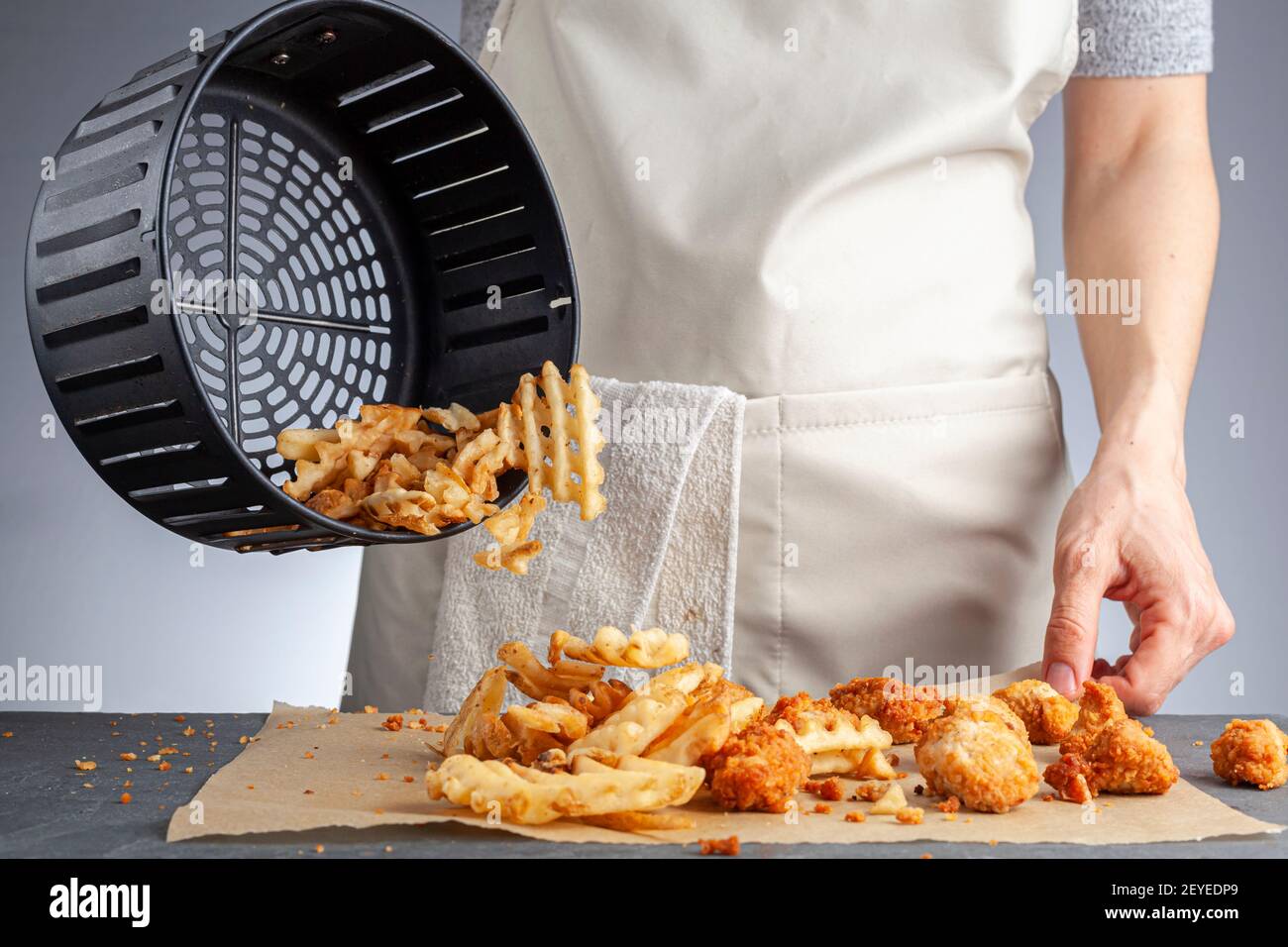 Eine Frau dumpt frisch zubereitete Kartoffelwaffelfritten aus dem Korb zusammen mit Hühnchen-Nuggets auf eine Arbeitsplatte. Sie frittierte sie in der Airfryer mit sehr Stockfoto