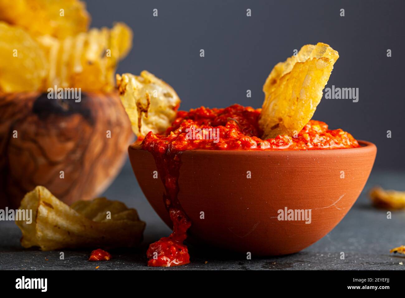 Eine Holzschüssel mit Kartoffelchips mit einer Terracotta-Schüssel mit Salsa oder scharfer Pfeffer-Dipping-Sauce. Ein Chips wurde eingetaucht. Etwas Durcheinander und verschüttet im Dunkeln Stockfoto