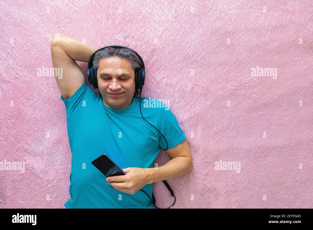 Der 50-jährige Mann hört Musik mit Kopfhörern zu Hause, auf dem Bett liegend. Entspannend, genießend. Stockfoto