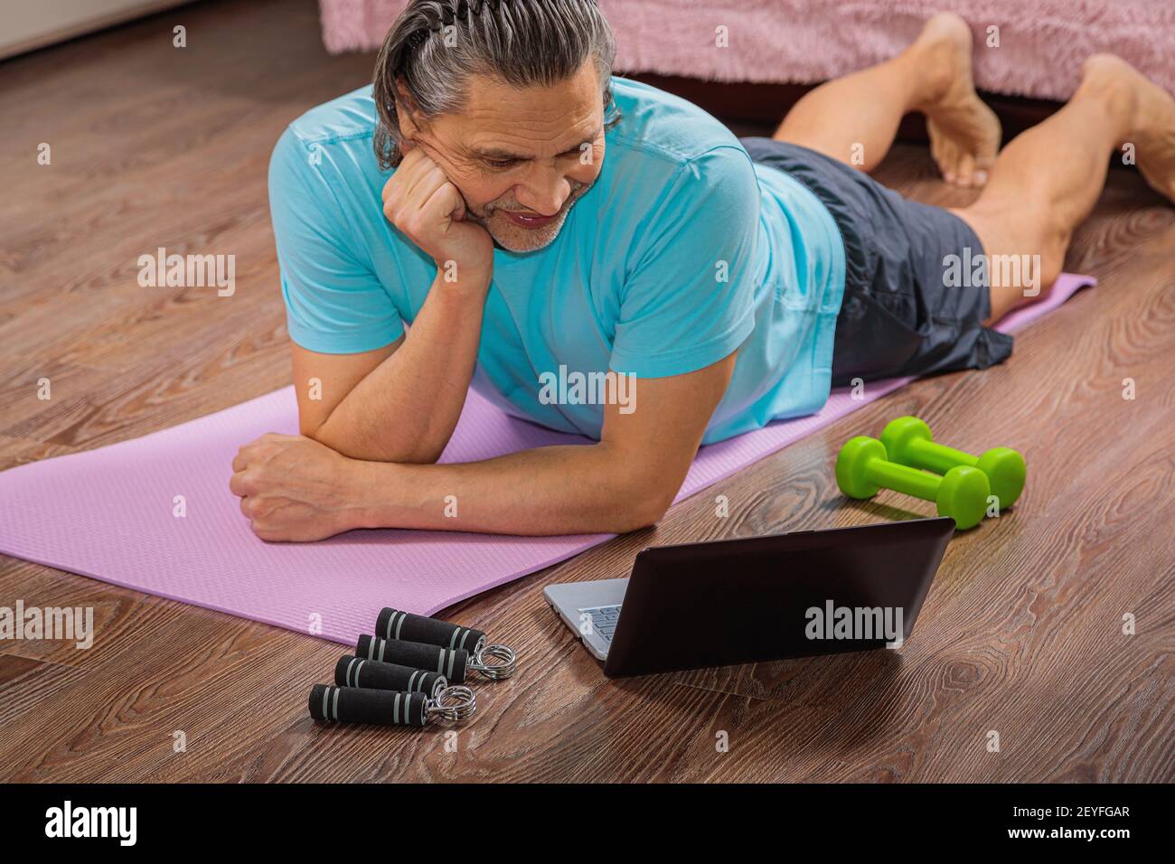 Der 50-jährige Mann führt Übungen durch, während er zu Hause auf dem Teppich liegt und den Computer anschaut. Während einer Pandemie trainiert eine Person in einer Wohnung über den Stockfoto