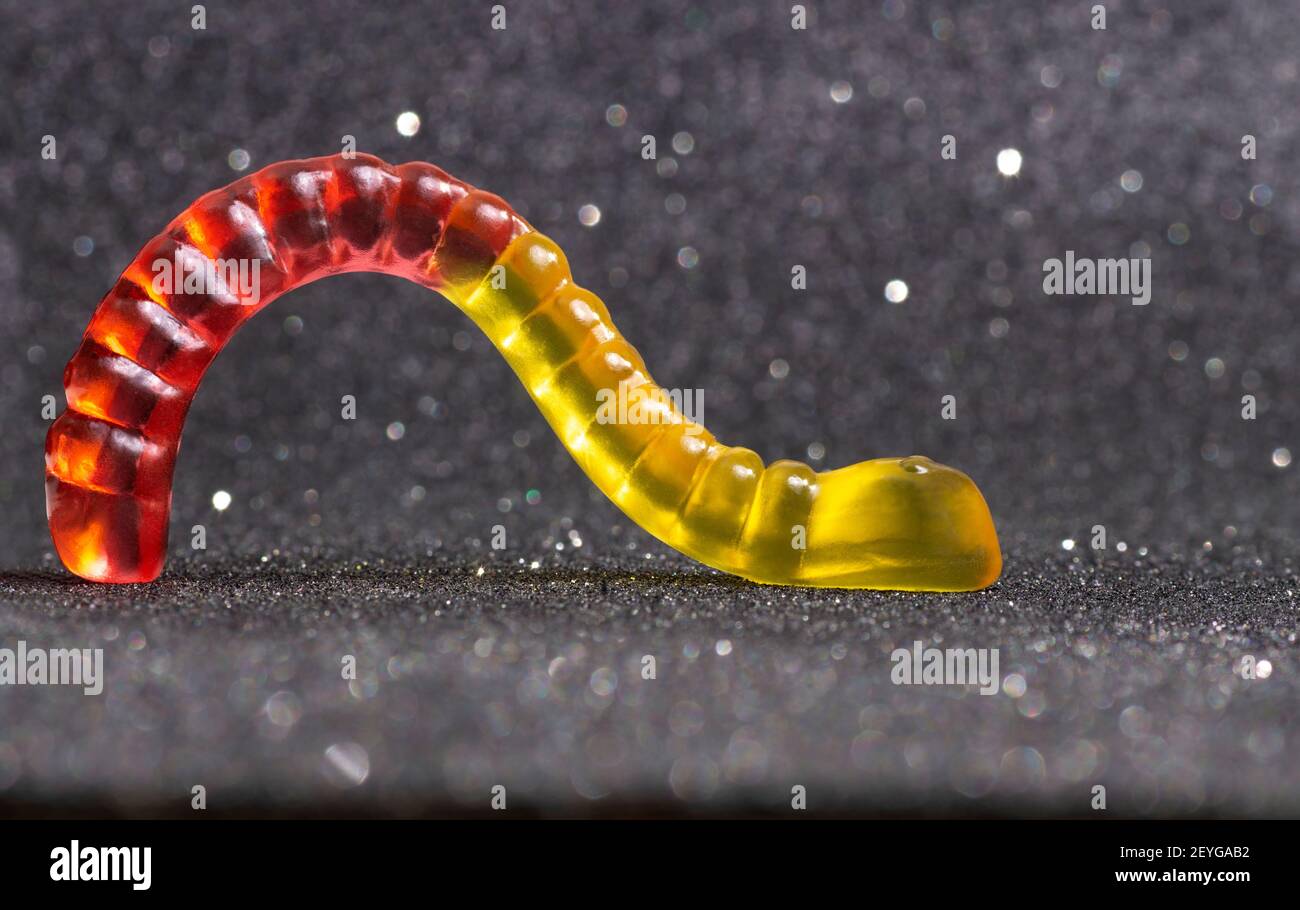 Nahaufnahme eines rot-gelben Gummiwurms mit Hintergrundbeleuchtung, der vor einem glitzernden Hintergrund kriecht. Stockfoto