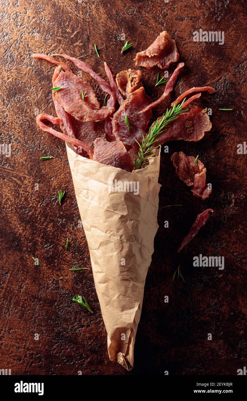 Scheiben von würzigem Trockenfleisch mit Rosmarin in einem Papierbeutel auf einem alten braunen Hintergrund. Draufsicht. Stockfoto