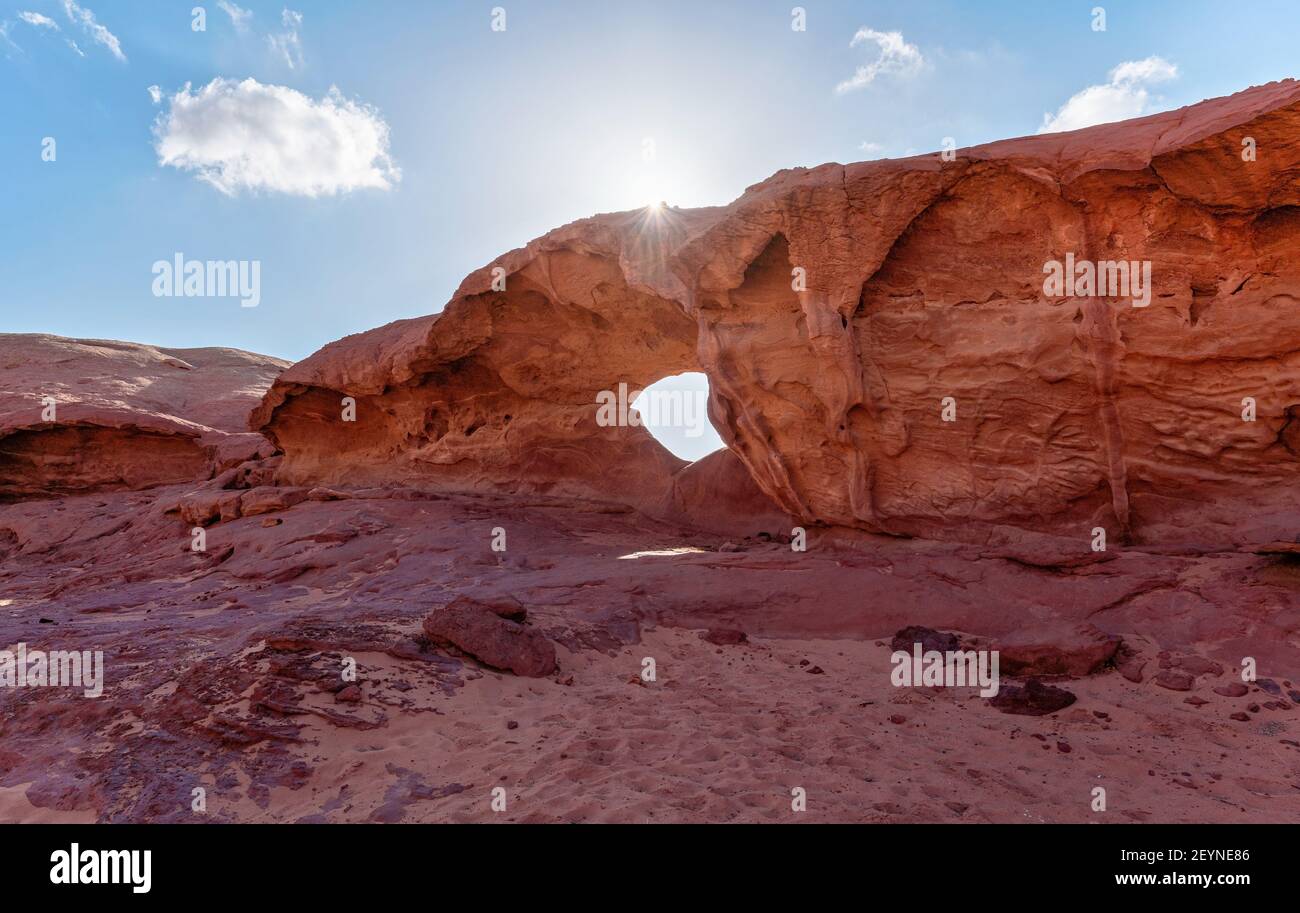 Kleiner Bogen oder kleine Felsenfensterformation in der Wadi Rum Wüste, helle Sonne scheint auf rotem Staub und Felsen, blauer Himmel darüber Stockfoto