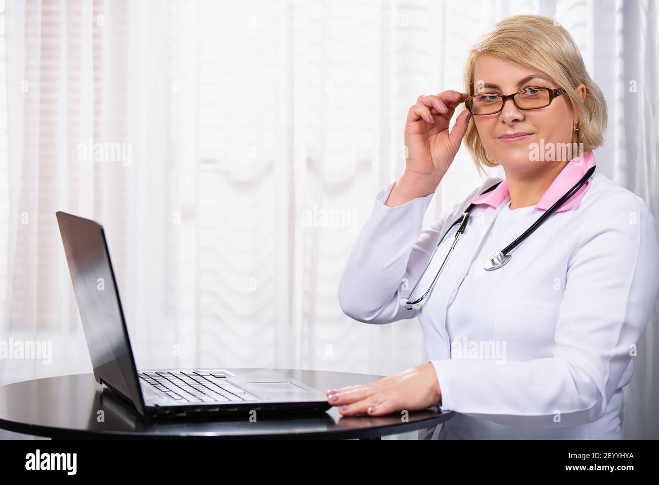 Ernsthafte Ärztin mit Laptop und Eingabe von Notizen in medizinische Zeitschrift sitzen am Schreibtisch. Ältere Frau, professioneller Arzt mit weißem Mantel Stockfoto