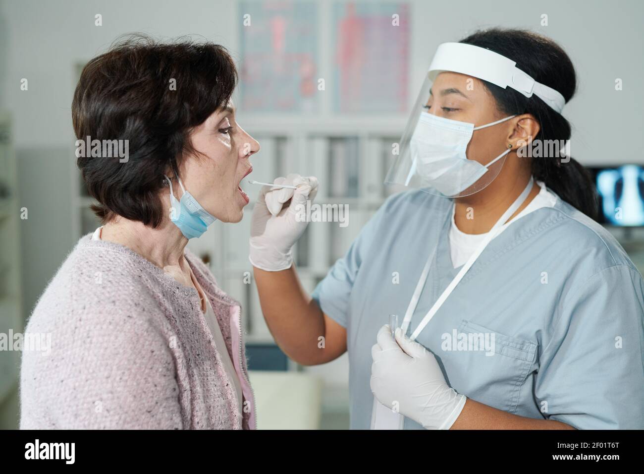 Reife Frau mit offenem Mund sitzt vor jungen Krankenschwester, die sie auf Kovid mit oralem Tupfer in medizinischen Tests testet Büro der zeitgenössischen Kliniken Stockfoto