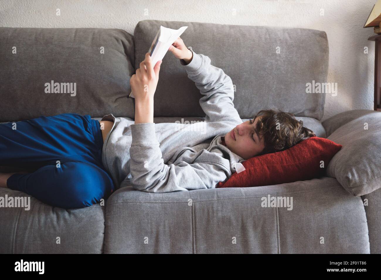 Junger Mann, der sich bequem auf der Couch ausdehnt und ein Buch liest. Mann, der sich zu Hause entspannt Stockfoto