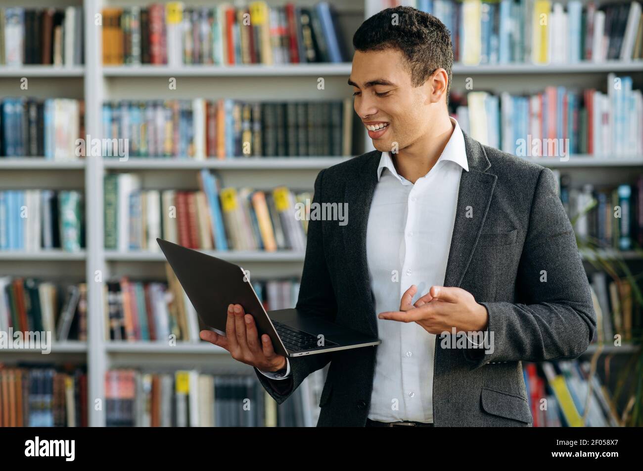 Erfolgreicher männlicher Angestellter benutzt Laptop, steht im Büro oder in der Bibliothek, sucht einige Informationen nach Projekt. Glücklicher hispanischer Geschäftsmann, der lächelnd auf den Laptop-Bildschirm schaut Stockfoto