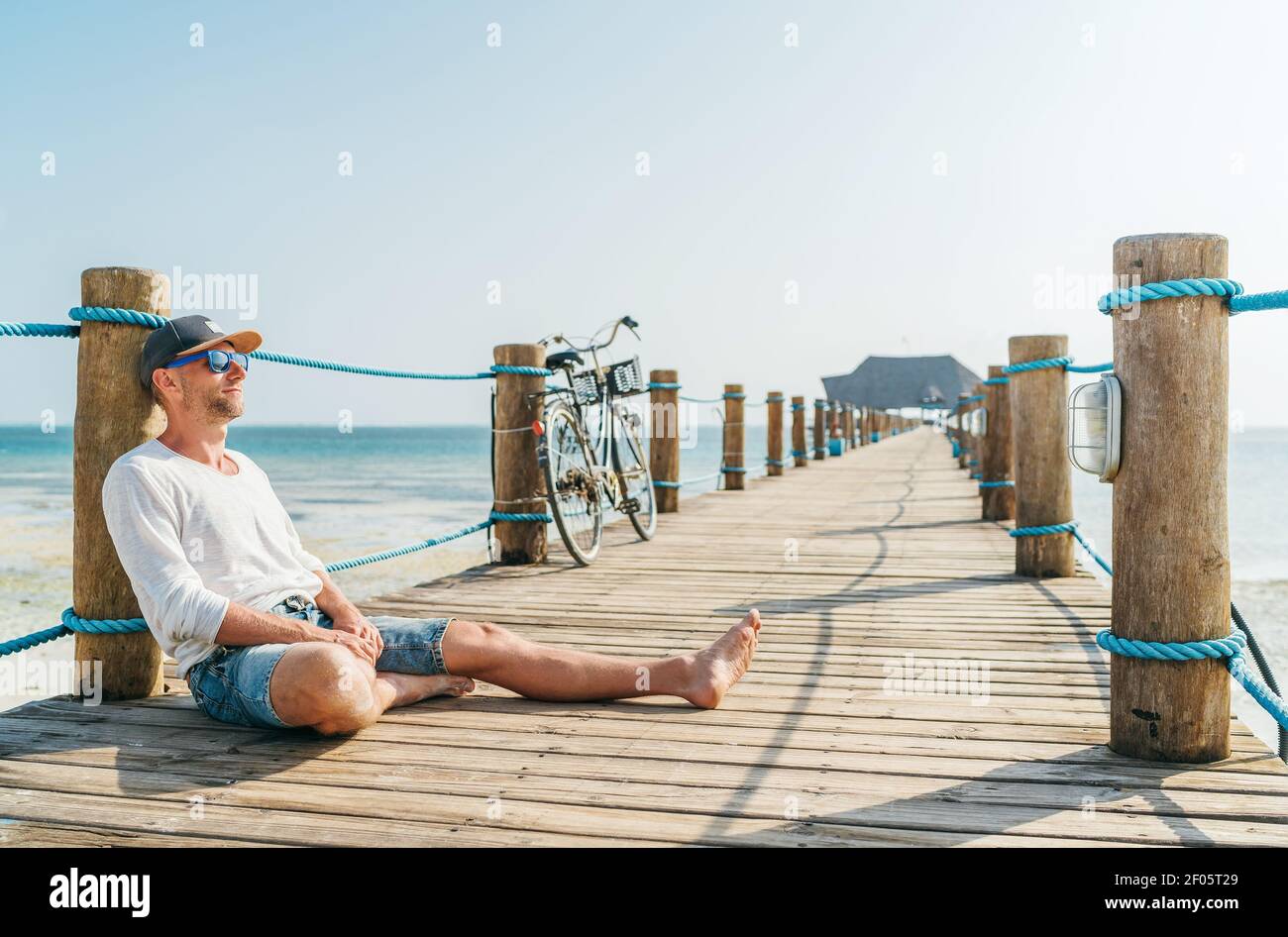 Porträt eines glücklichen lächelnden Mannes in leichten Sommerkleidung und Sonnenbrille sitzen und genießen Zeit auf hölzernen Seebrücke. Sorgloser Urlaub in Tropf Stockfoto