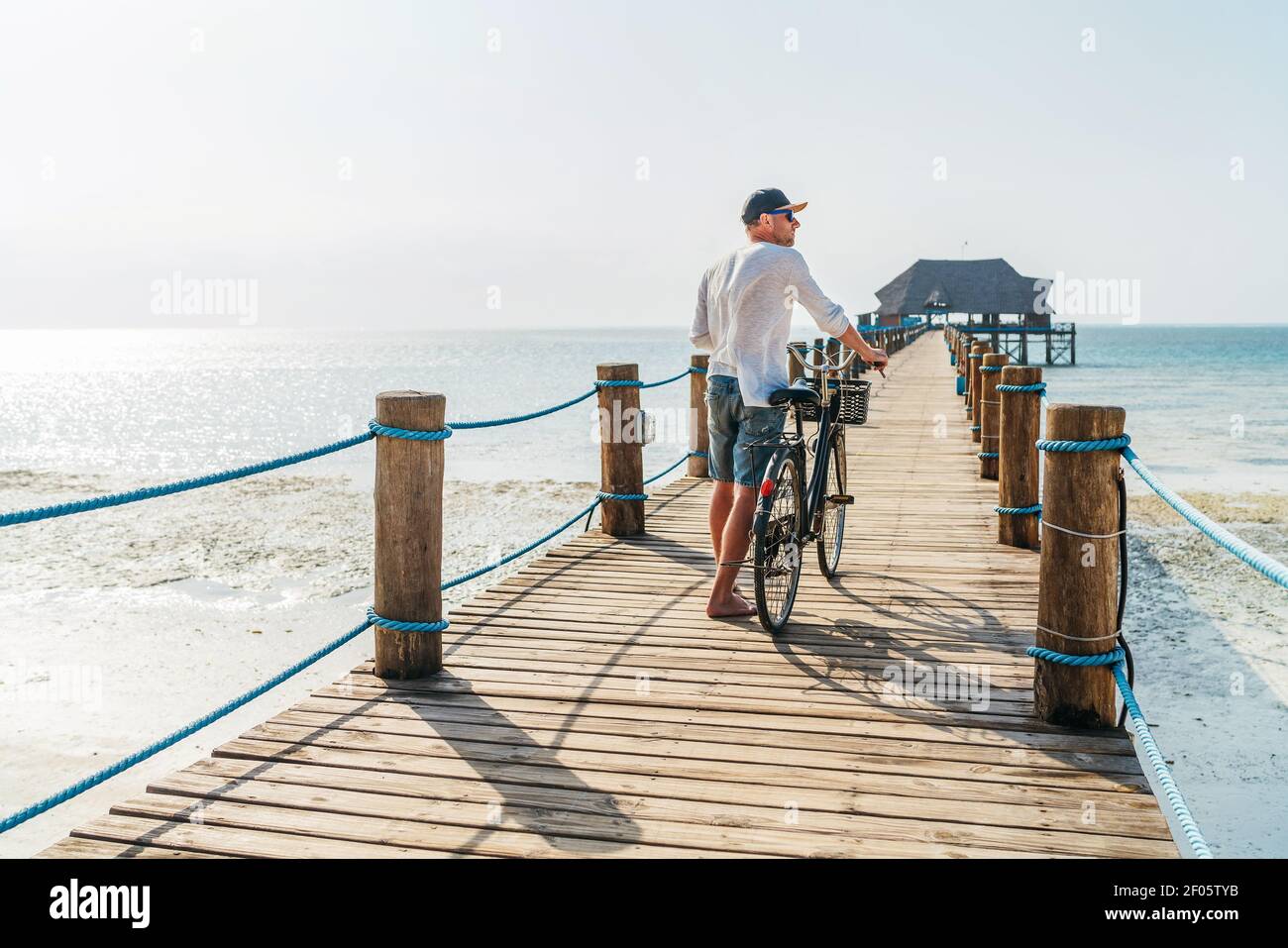 Rückansicht Porträt eines Mannes in leichte Sommerkleidung gekleidet, Sonnenbrille mit altem Fahrrad auf dem hölzernen Seebrücke auf dem sandigen Sansibar Strand.sorglos V Stockfoto