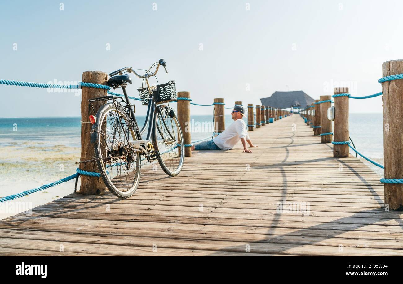 Altes Fahrrad auf dem hölzernen Seebrücke und ein glücklicher lächelnder Mann, der in leichten Sommerkleidung gekleidet ist, sitzt und die Zeit im Hintergrund genießt. Sorgloser Urlaub in t Stockfoto