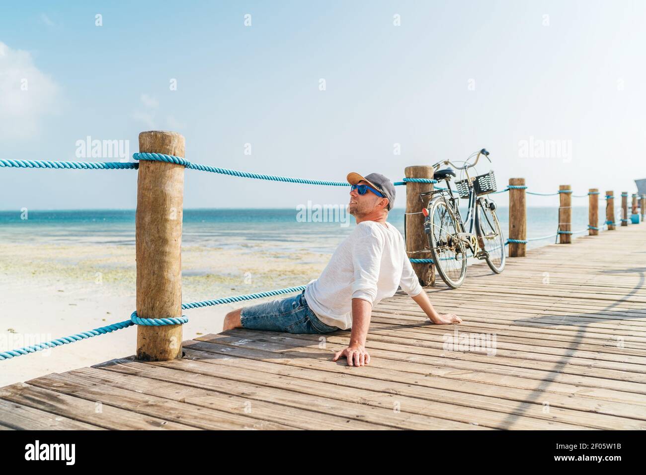 Portrait von entspannenden Mann in leichten Sommerkleidung gekleidet und Sonnenbrillen sitzen und genießen Sie Zeit und Blick auf den Strand auf Holz pier. Sorgloser Urlaub in t Stockfoto