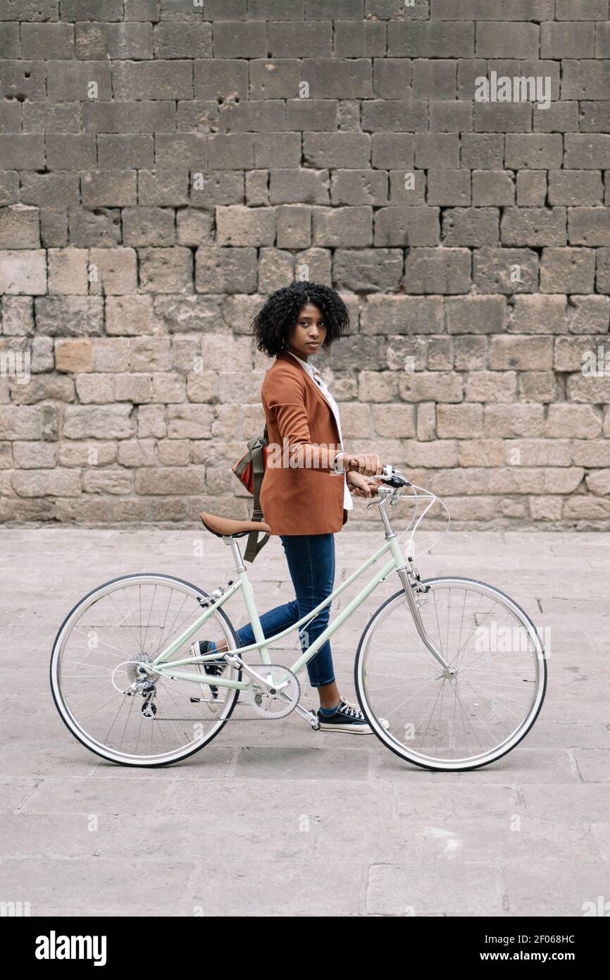 Seitenansicht einer schwarzen Frau in elegantem Casual-Stil, die mit dem Fahrrad auf der Straße läuft und die Kamera anschaut Stockfoto