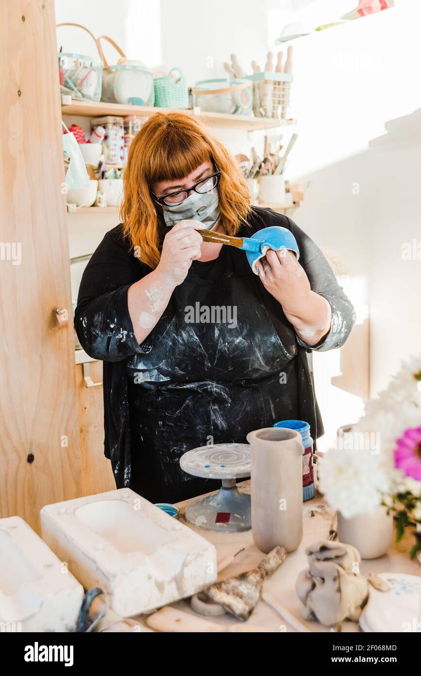 Nicht erkennbare Frau in Maske und schmutzige schwarze Schürze Stehtisch mit Töpferscheibe und Tonvase in der Nähe von Formen für Keramik während der Malerei handgefertigten Cera Stockfoto
