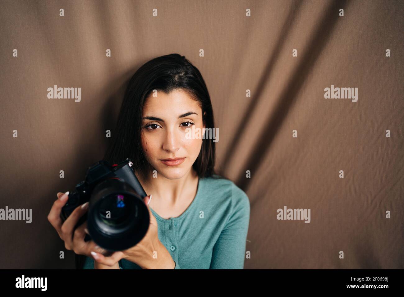 Millennial weiblich mit professioneller Fotokamera sitzt auf braun zerknittert Textiler Hintergrund und Blick auf die Kamera zu Hause Stockfoto