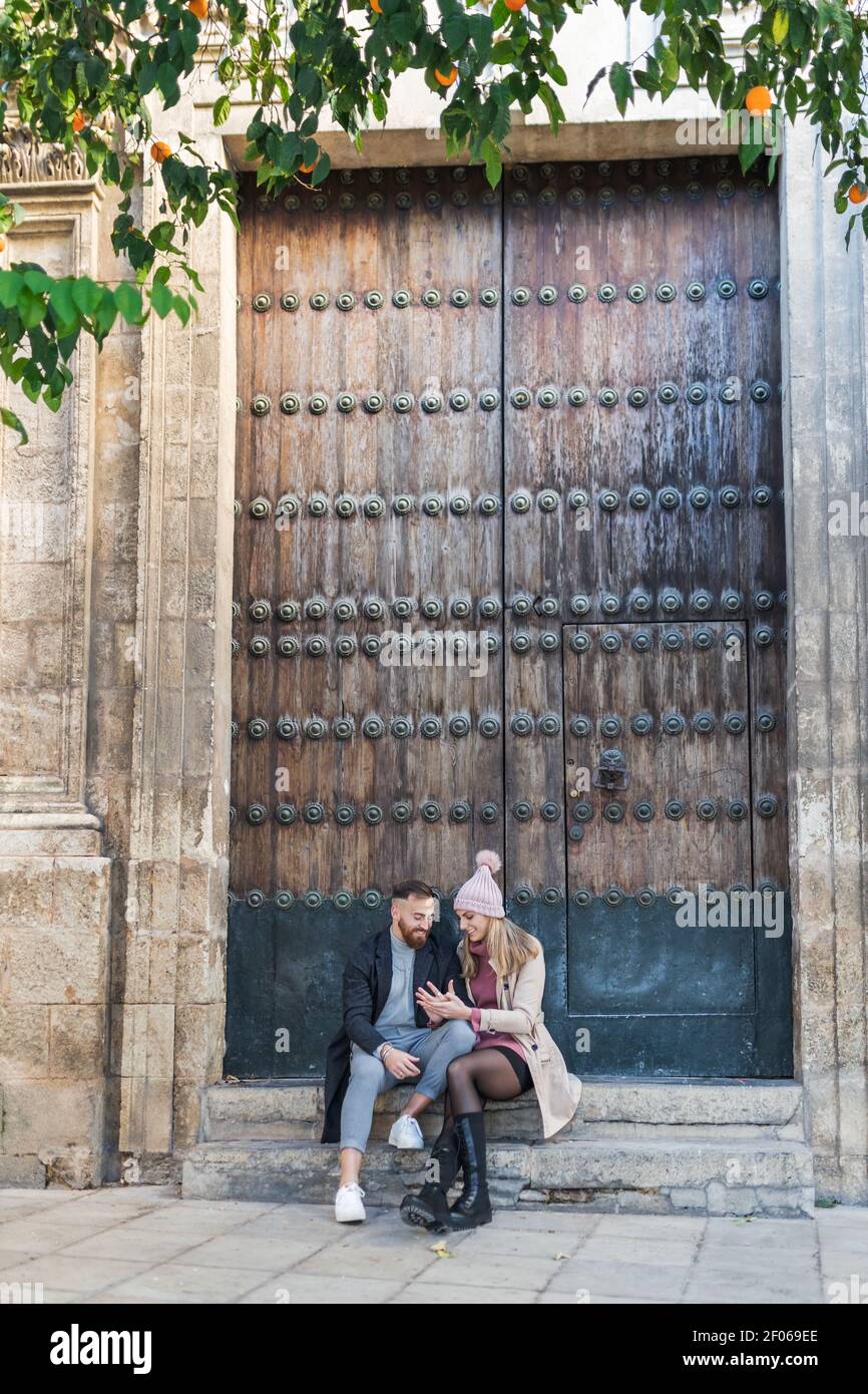 Liebevolles Paar in stilvoller Oberbekleidung sitzen neben riesigen Türen und Hände halten, während Sie das Wochenende in der Stadt gemeinsam genießen Stockfoto