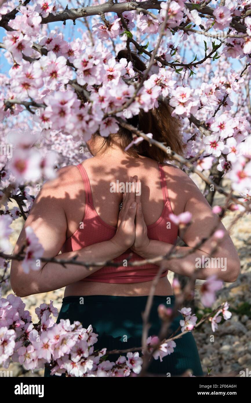 Anonyme Frau im aktiven Ohr stehend in Yoga-Pose mit Namaste Hände hinter dem Rücken, während Sie Achtsamkeit unter blühenden Mandelbäumen üben Stockfoto