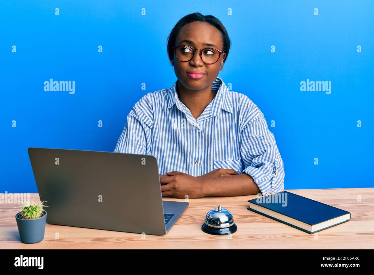 Junge afrikanerin, die an der Hotelrezeption mit einem Laptop arbeitet, lächelt und schaut zur Seite und starrt wegdenkend. Stockfoto