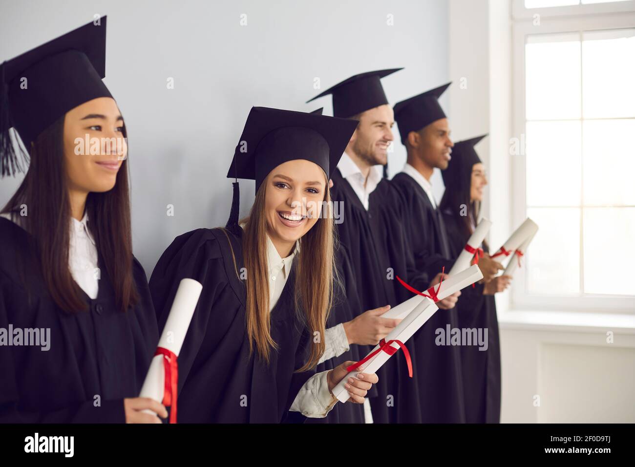 Frau mit einem Diplom in der Hand steht in einer Reihe in der Nähe seiner multiethnischen Klassenkameraden Freunde in der Nähe der Wand. Stockfoto