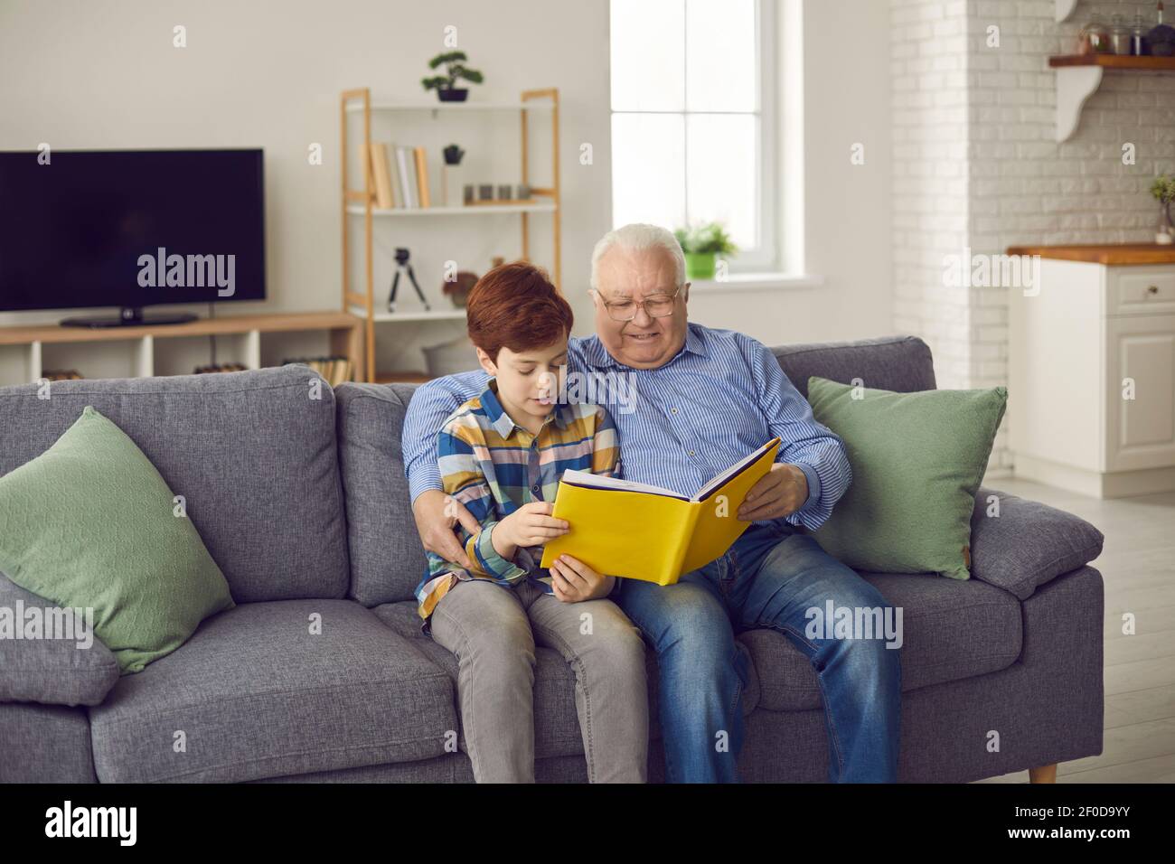 Glücklicher Großvater liest seinem Enkel ein Buch, während er darauf sitzt Sofa im Wohnzimmer Stockfoto