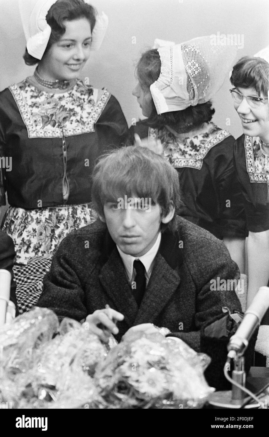 Holländische Mädchen stehen hinter Lead-Gitarrist George Harrison auf einer Pressekonferenz am Flughafen Schiphol, als die Beatles am 5. Juni 1964 in Amsterdam, Holland, ankamen. Stockfoto