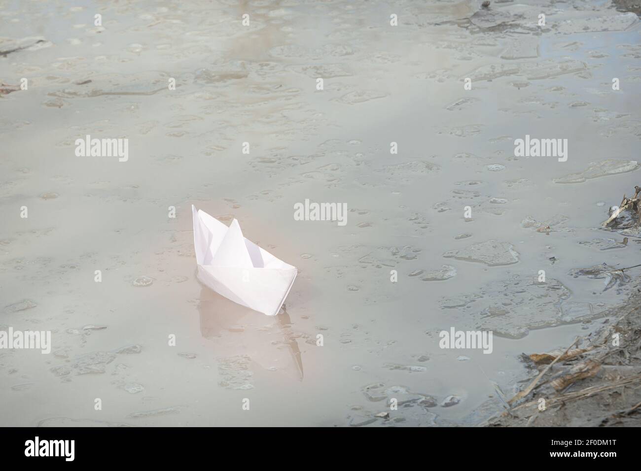 Weißes handgemachtes Papierboot segelt zum Ufer. Schmutziges Wasser mit Eisschollen. Frühfrühling Konzept, Freiheit. Stockfoto