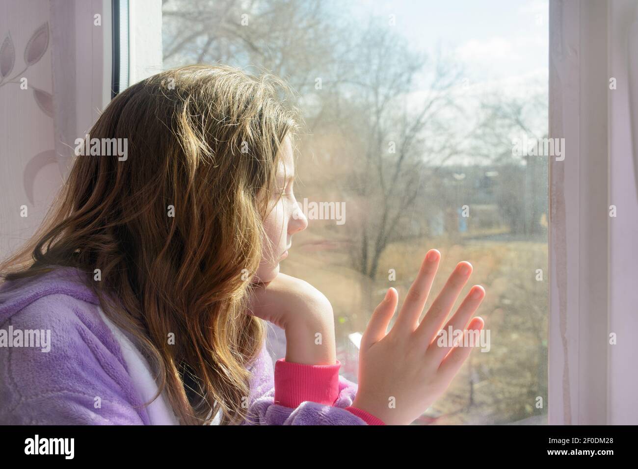 Das Mädchen im Teenageralter schaut morgens aus dem Fenster. Du kannst nicht gehen, traurige Emotionen. Nahaufnahme, Seitenansicht. Stockfoto