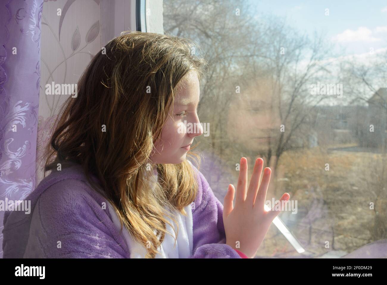 Teenager-Mädchen schaut aus dem Fenster bei Sonnenaufgang in der Sonne. Hand auf Glas. Nahaufnahme, Seitenansicht, selektiver Fokus. Stockfoto