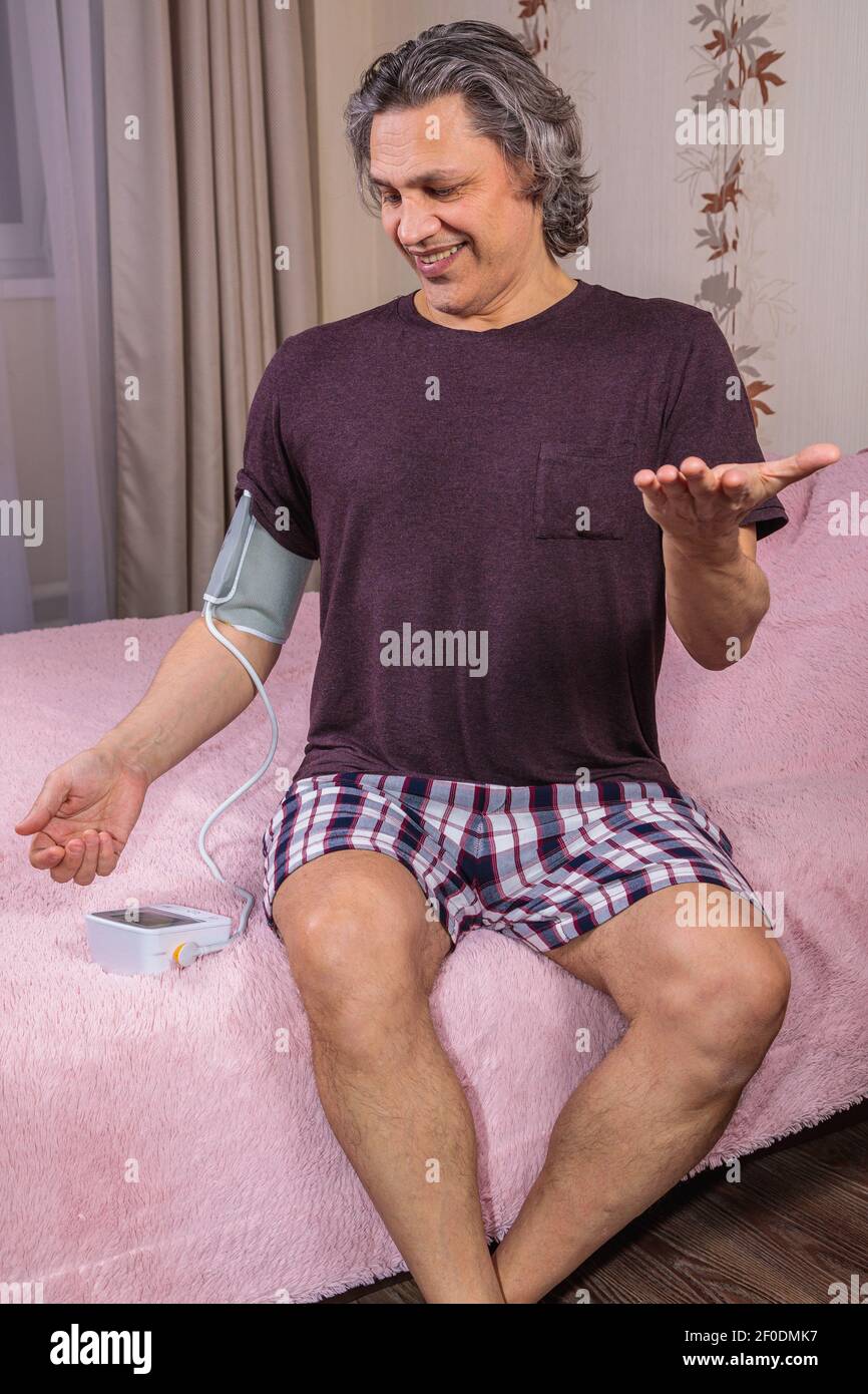 Ein 50-jähriger Mann misst seinen Blutdruck mit einem Blutdruck-Monitor zu Hause und sitzt auf seinem Bett in seiner Hauskleidung. Überrascht von der Einstutze Stockfoto