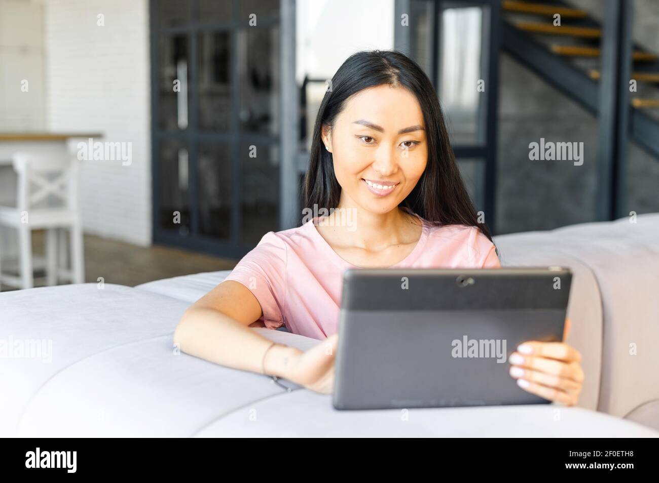 Fröhliche junge gut aussehende asiatische Frau verbringt Freizeit mit einem digitalen Tablet zu Hause, eine Frau schaut Filme, chatten in sozialen Netzwerken Stockfoto