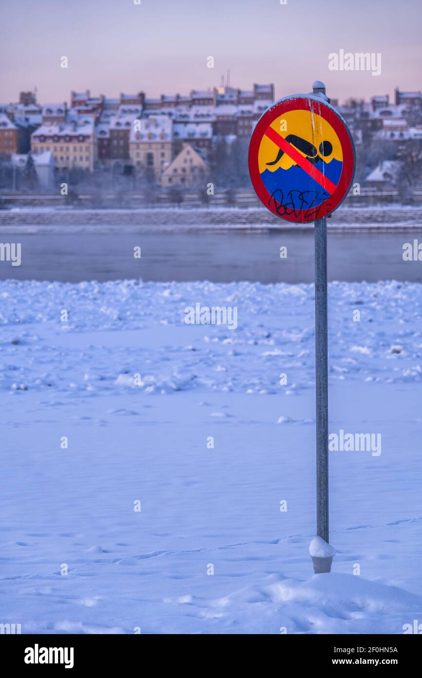 Kein Schwimmwarnschild im Schnee an einem Strand am Wisla Fluss im Winter, Stadt Warschau bei Sonnenaufgang in Polen, Altstadt Skyline im Hintergrund. Stockfoto