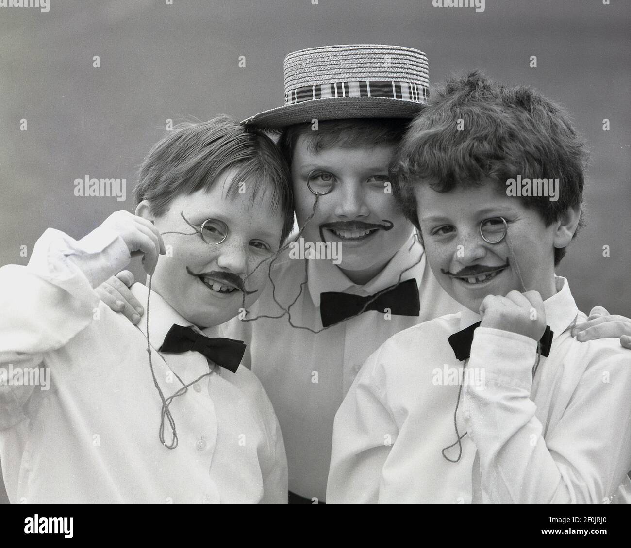 1980s, historisch, drei junge Jungen in ausgefallener Kleidung für ein Schulspiel, mit bemalten Schnurrbärten, einem kleinen Augenglas oder Monocle und einem mit Strohhut oder Bootsfahrer. Sie könnten nur allgemeine Musiksaalfiguren sein oder vielleicht das Bild des belgischen Detektivs Hercule Poirot, einer beliebten fiktiven Figur der berühmten Schriftstellerin Agatha Christie, einschüchtern. Stockfoto