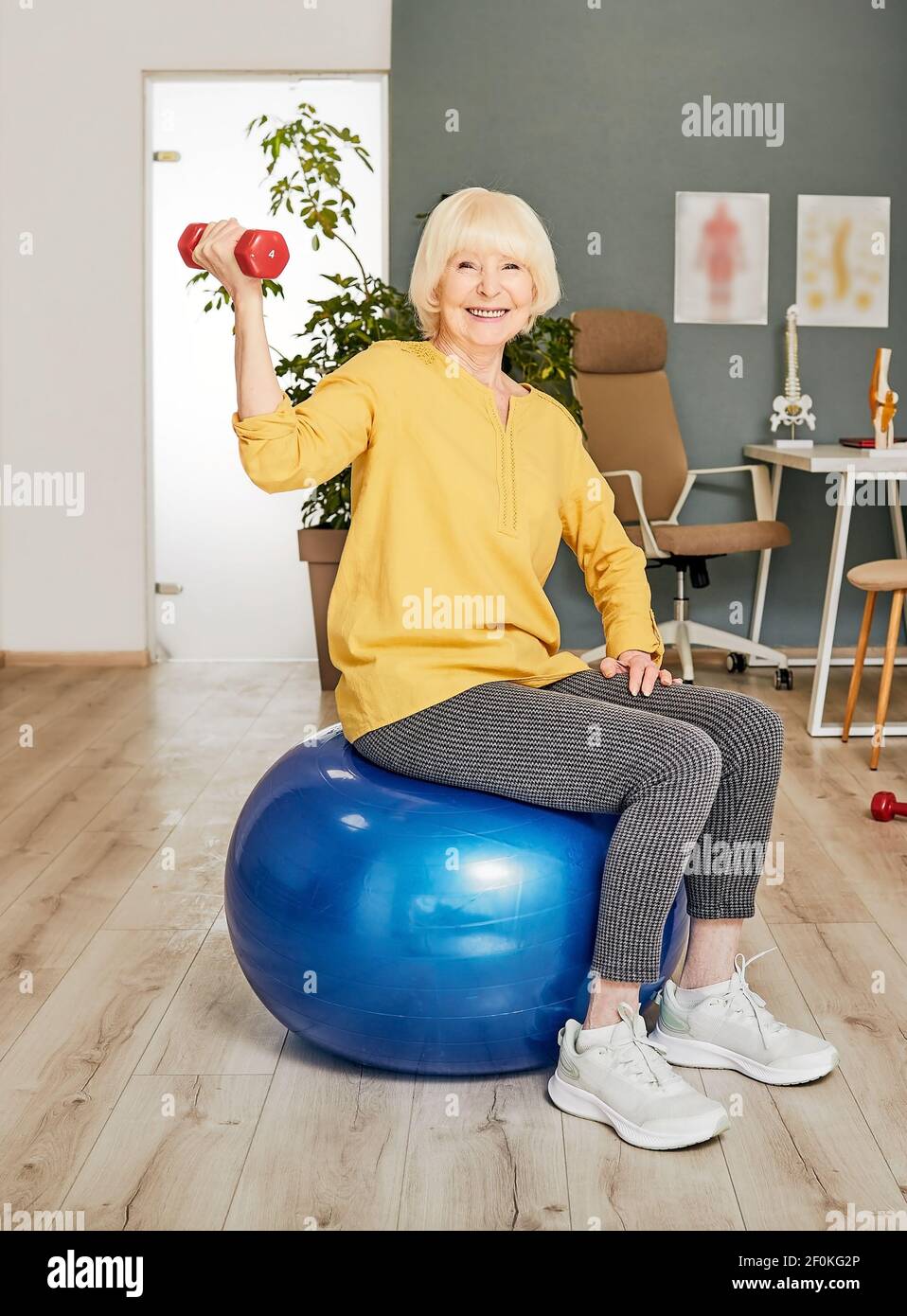 Ältere Frau mit einer Hantel in der Hand sitzt auf einem Fitnessball während der Bewegungstherapie in einem Rehabilitationszentrum Stockfoto