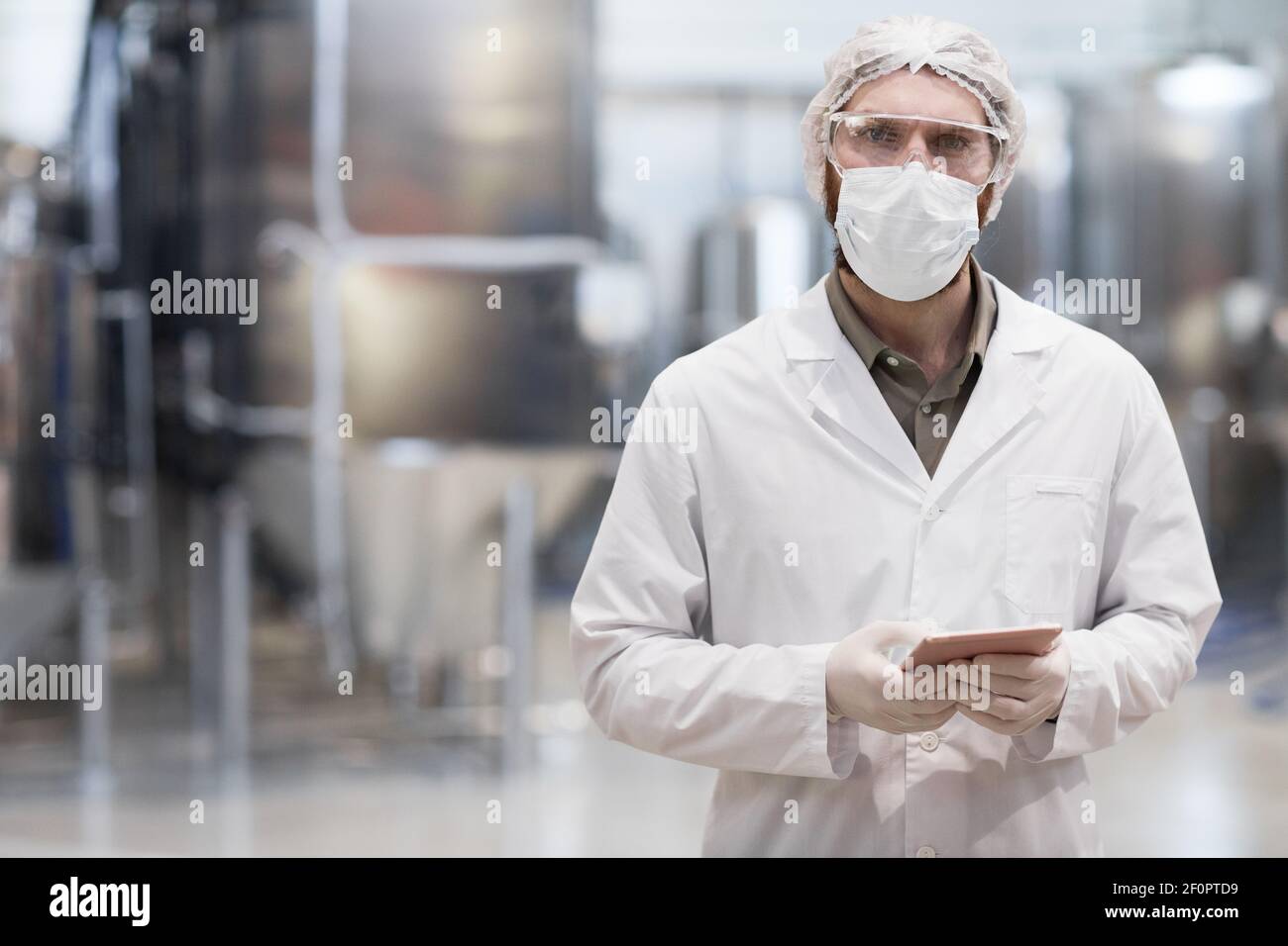 Waist-up-Porträt eines erwachsenen Mannes, der in einer chemischen Fabrik arbeitet und die Kamera anschaut, während er einen schützenden Labormantel und eine Maske trägt, Platz kopieren Stockfoto