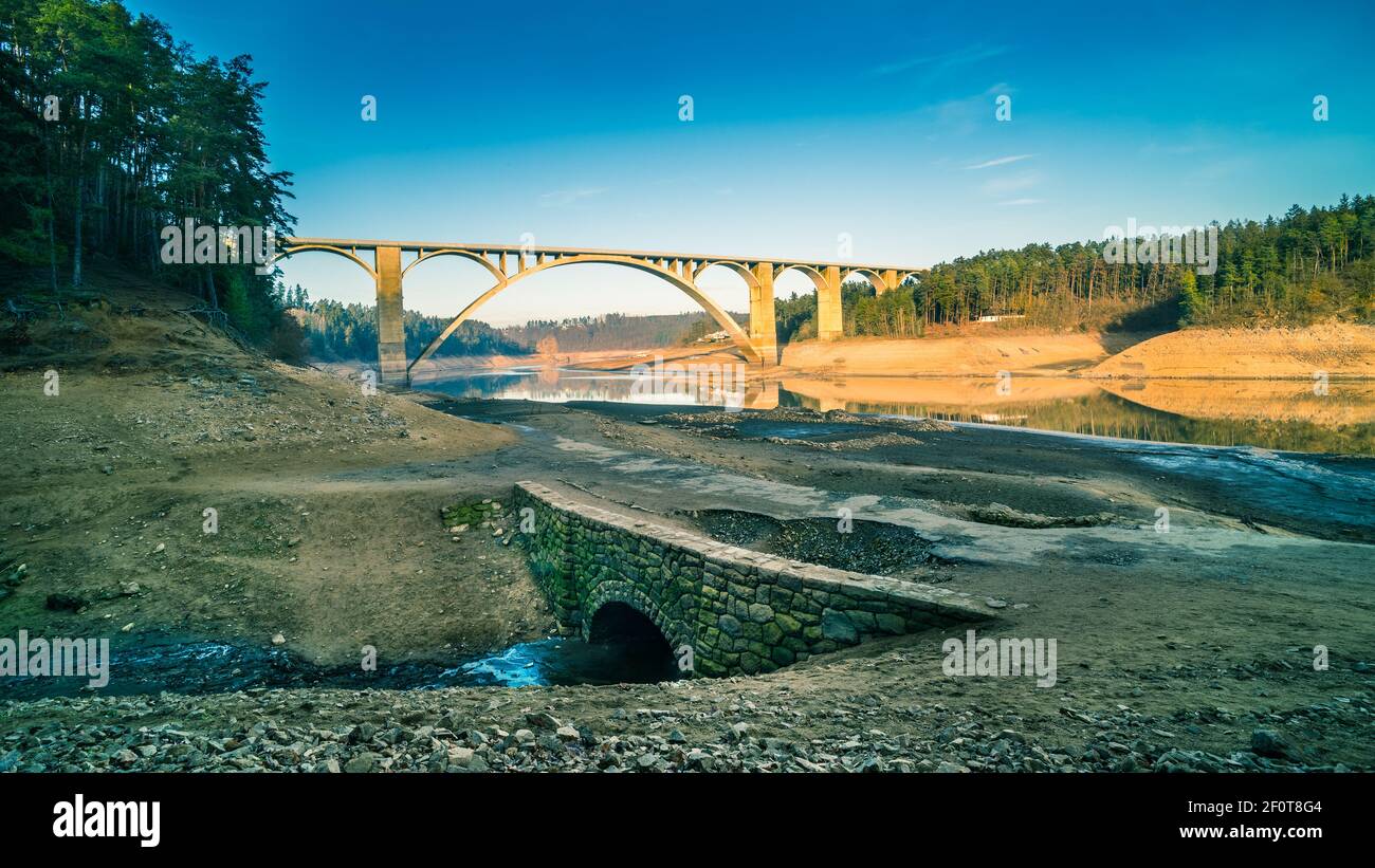Hoch gewölbter Viadukt über leeren Orlik Damm und original Moldau Flussbett. Kontrast von großem Beton und kleiner alter Steinstraßebrücke. Podolsko, Tschechien. Stockfoto