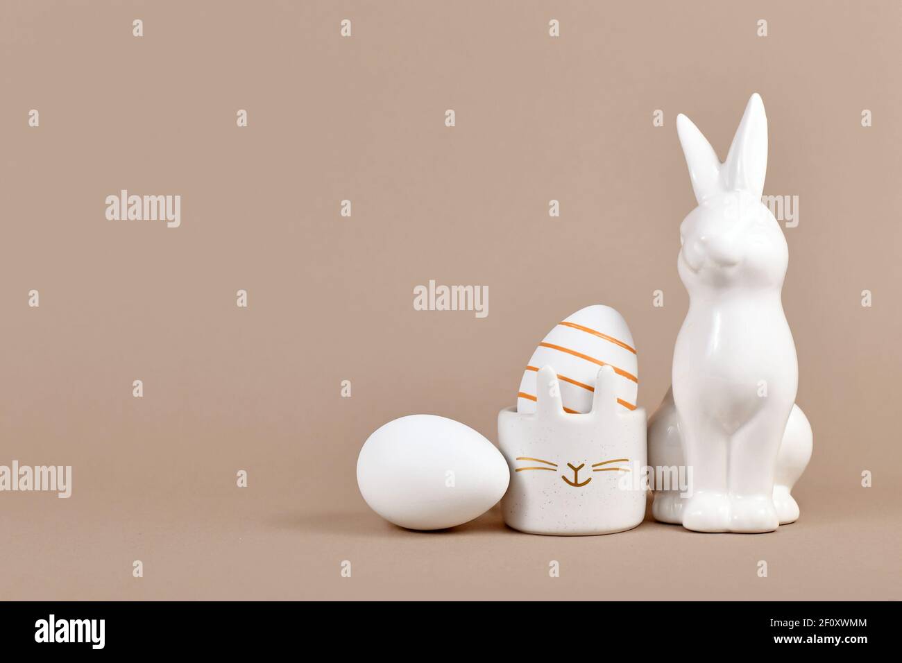Goldgestreiftes Osterei in Hase Eierbecher daneben Hasenfigur und weißes Ei auf der Seite des beigen Hintergrunds Mit leerem Kopierbereich Stockfoto