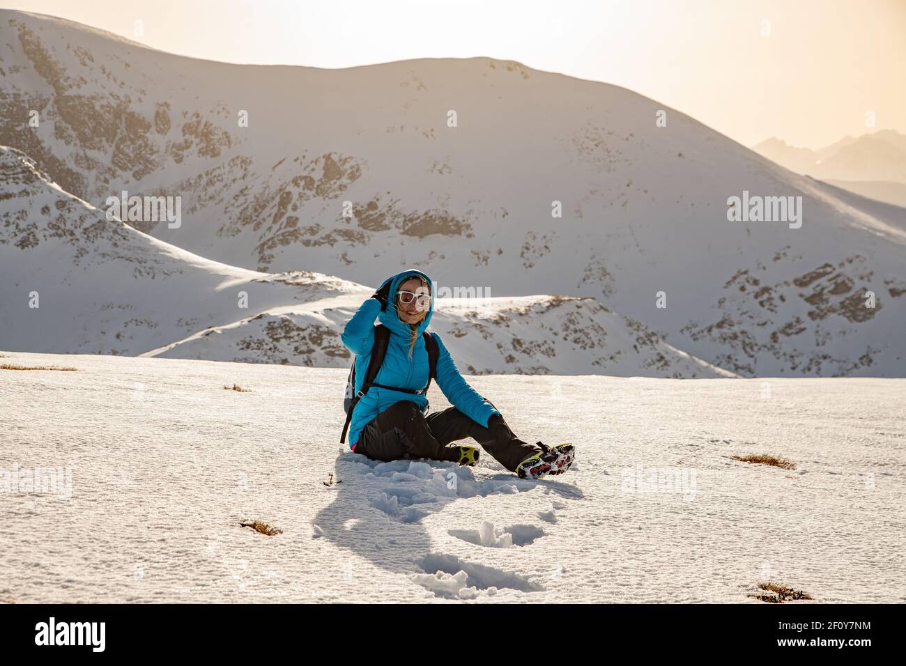 Blonde Tourist in Sonnenbrille sitzt auf dem Schnee in den Bergen. Stockfoto